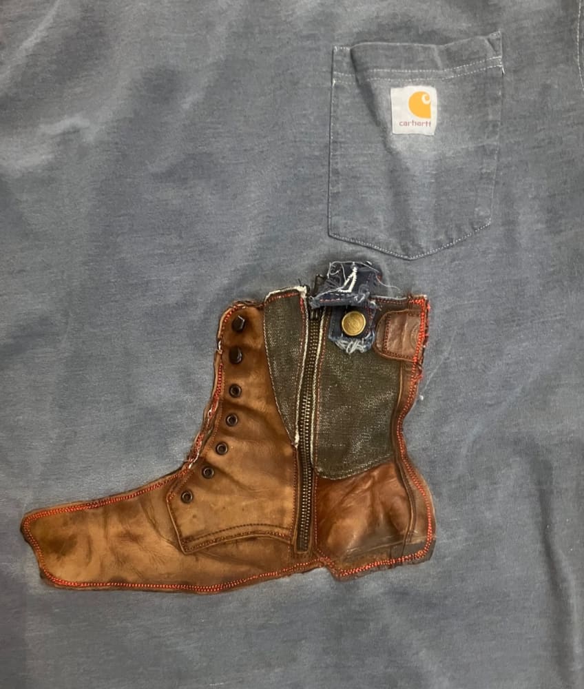 A grey Carhartt t-shirt with a brown and green boot appliqué featuring laces, a zipper, and a button.