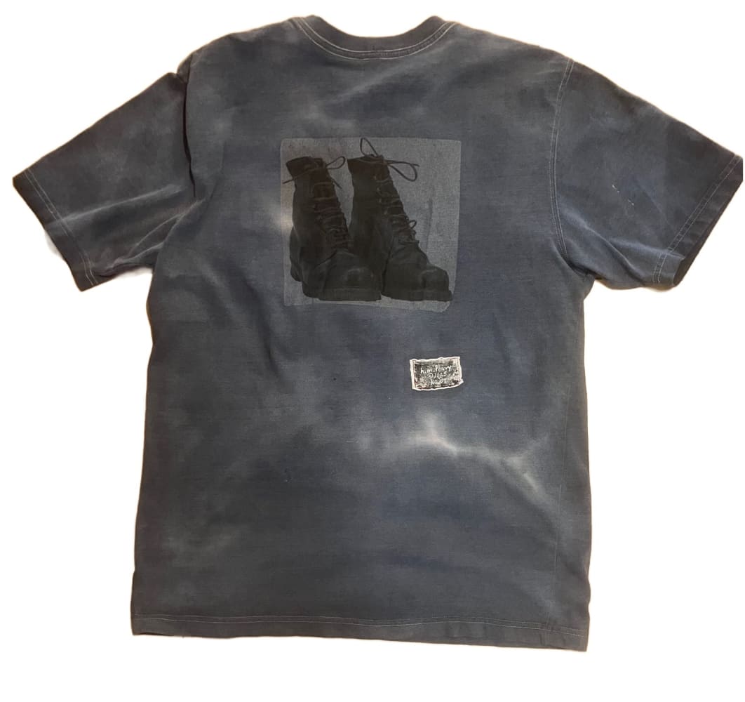A distressed, dark grey tie-dye t-shirt with a black graphic print of two combat boots on the back, and a small, rectangular woven label below it.