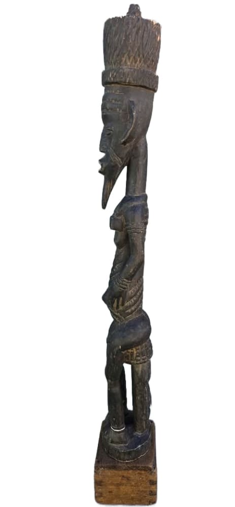 A tall, dark wooden African tribal sculpture of a standing figure with a prominent headdress, beard, and intricate carvings.