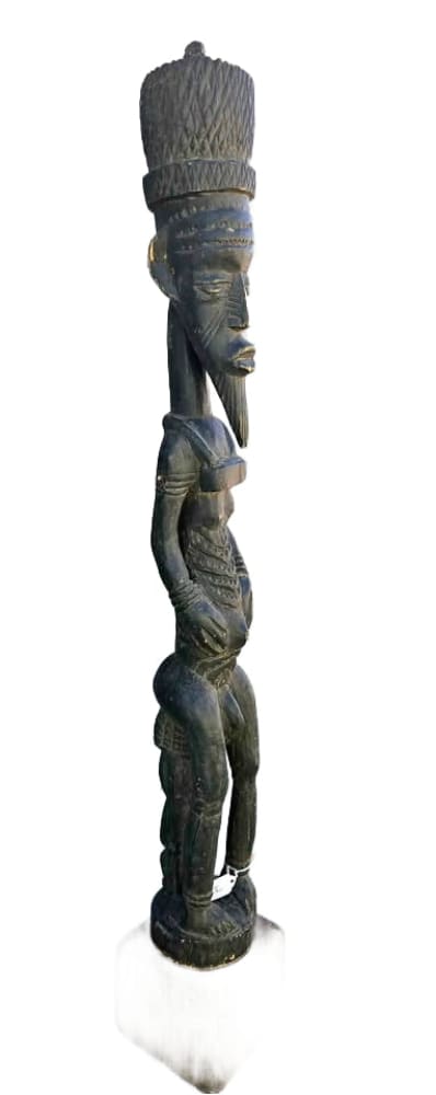 A tall, dark wooden African fertility statue depicting a figure with a conical headdress, elongated neck, and prominent stomach.