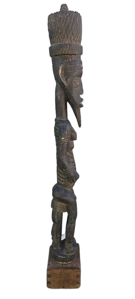Tall, dark, carved wooden tribal statue of a standing figure with a large headdress and pointed beard.