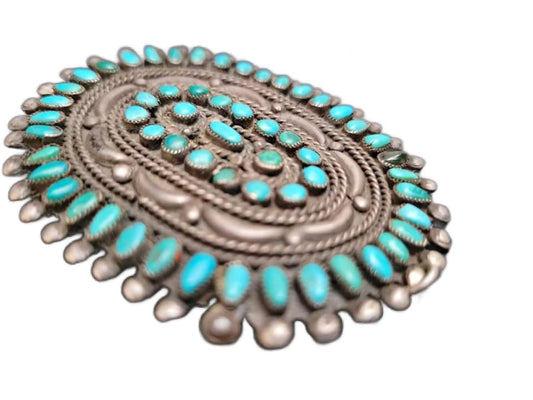 An oval silver belt buckle adorned with numerous turquoise stones.