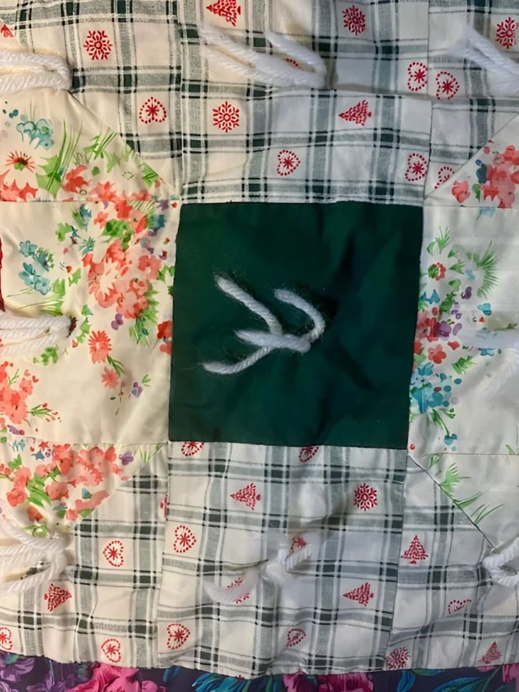 A handmade quilt with diverse fabric patterns, including floral, plaid with holiday motifs, and a solid dark green square, tied together with white yarn.