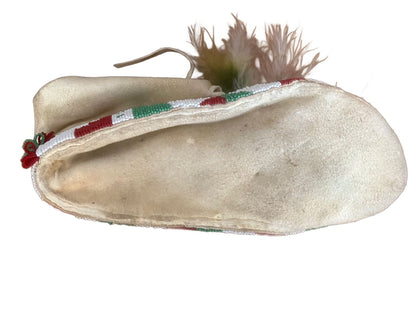 A single, light-colored, beaded moccasin with feather embellishments.