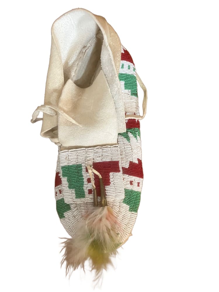 A single, handmade white moccasin with red and green beadwork and feather adornments.