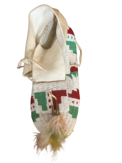 A single, handmade white moccasin with red and green beadwork and feather adornments.