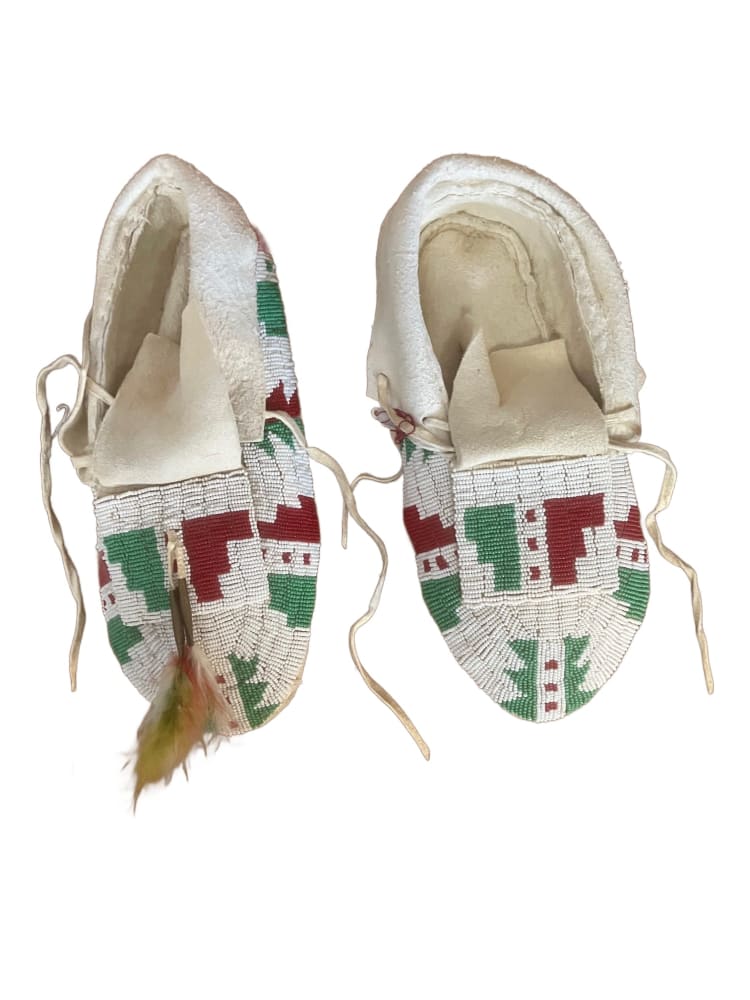A pair of white, green, and red beaded moccasins with a small feather accent on one.