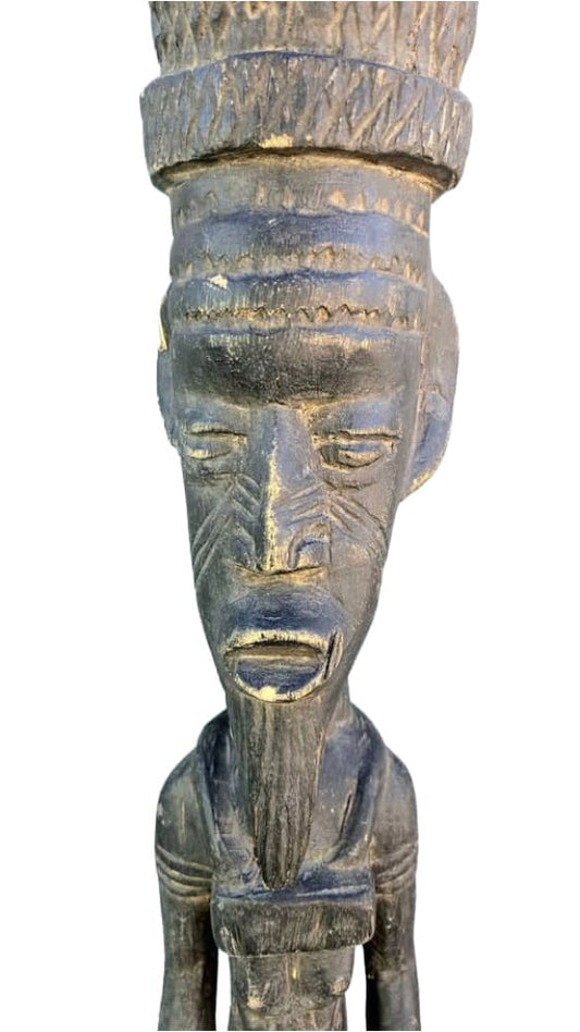 A weathered, dark wooden African tribal sculpture depicting a stylized human figure with a headdress, facial scarification, and a long beard.