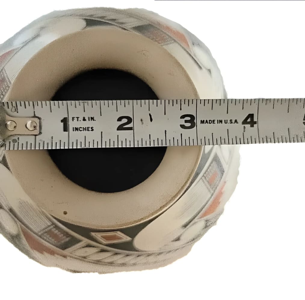 A white ceramic pot with intricate black and red geometric patterns, measured with a tape measure across its opening.
